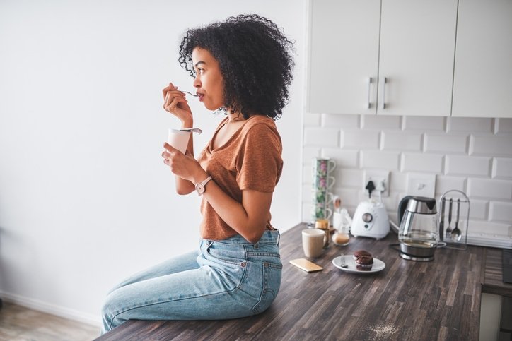 Imagem mostra mulher sentada em cima de bancada de cozinha comendo iogurte - Metrópoles