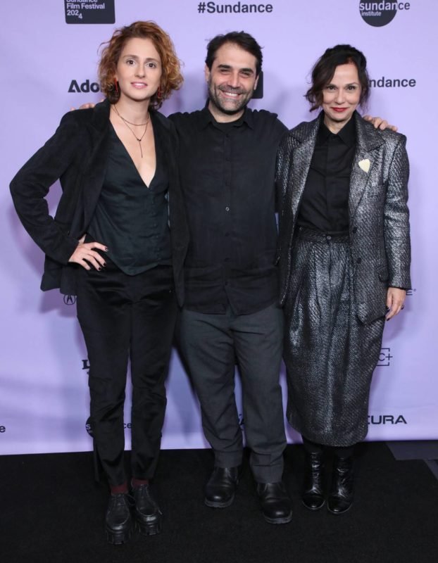 Três pessoas vestidas de preto em frente a um painel lilás