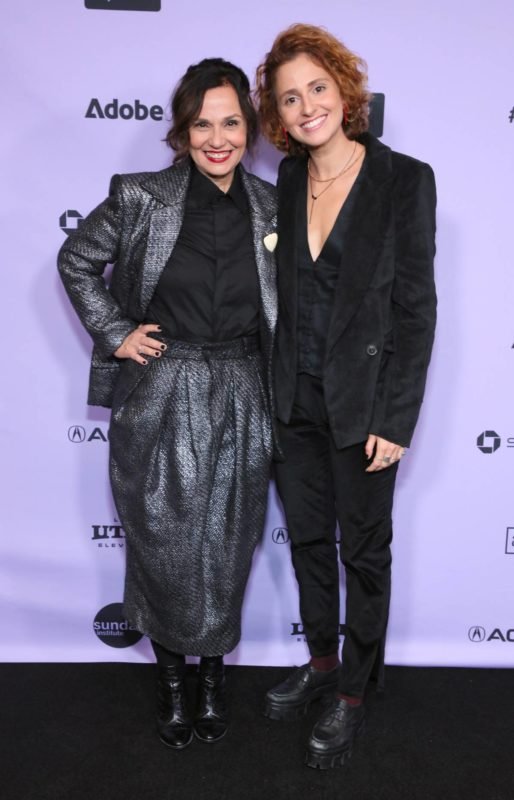 Duas mulheres com roupa preta em frente a um painel lilás