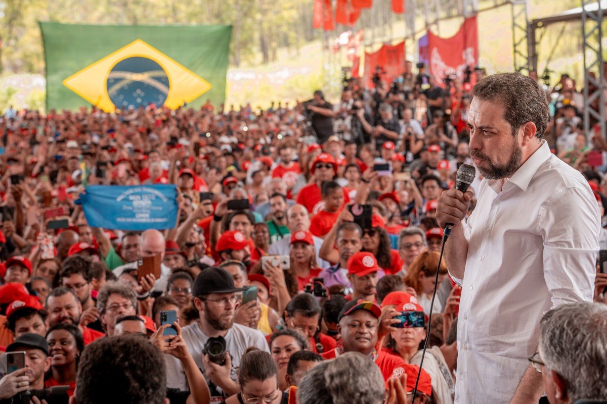 Imagem colorida mostra Guilherme Boulos, homem branco de cabelo e barba castanhos, de camisa branca, falando em um palco. No fundo, uma multidão de pessoas vestidas de roupas vermehas - Metrópoles