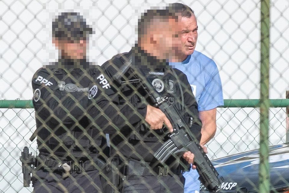 Marcola, homem branco, magro, de cabelos curtos e escuros, veste uniforme azul, usado na prisão; ele é escoltado por policiais de uniformes pretos e com rostos borrados