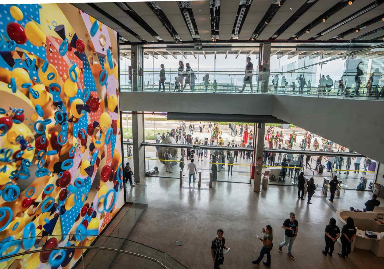 Fotografia colorida de um interior de um museu com pessoas participando de atividades culturais